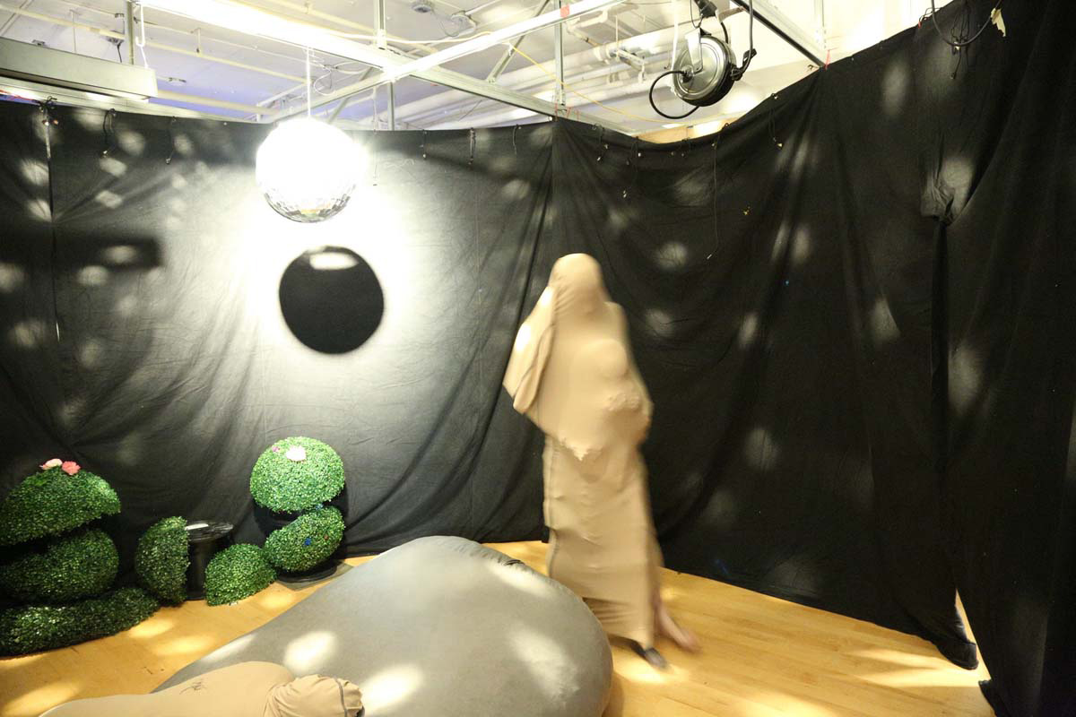 2 people with nylon covering their bodies in a black curtained room with lights from a disco ball, beanbag chair and plants 